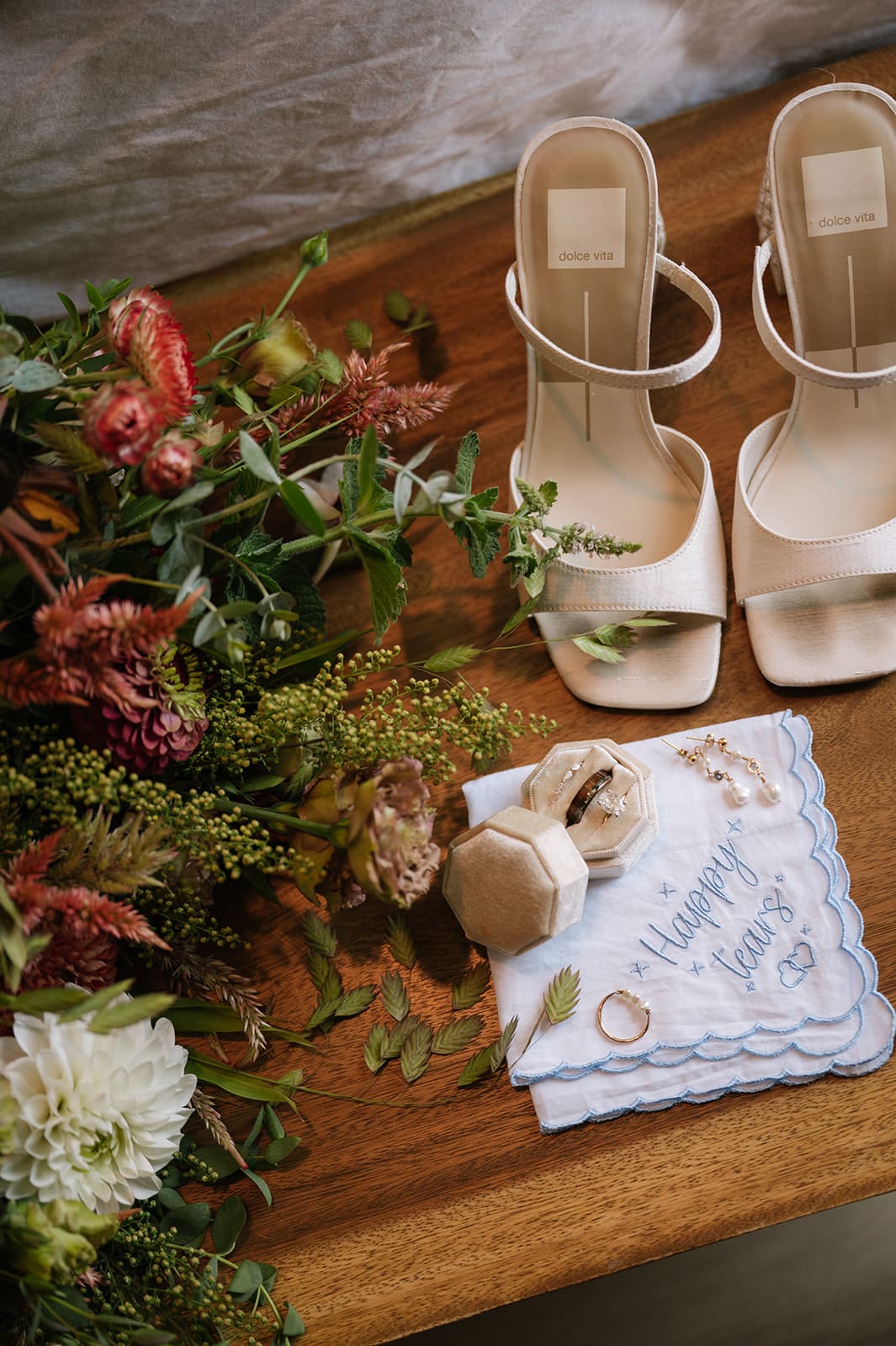A pair of beige high-heeled sandals, a blue embroidered handkerchief with wedding rings, a jewelry box, and a bouquet of flowers arranged on a wooden surface.