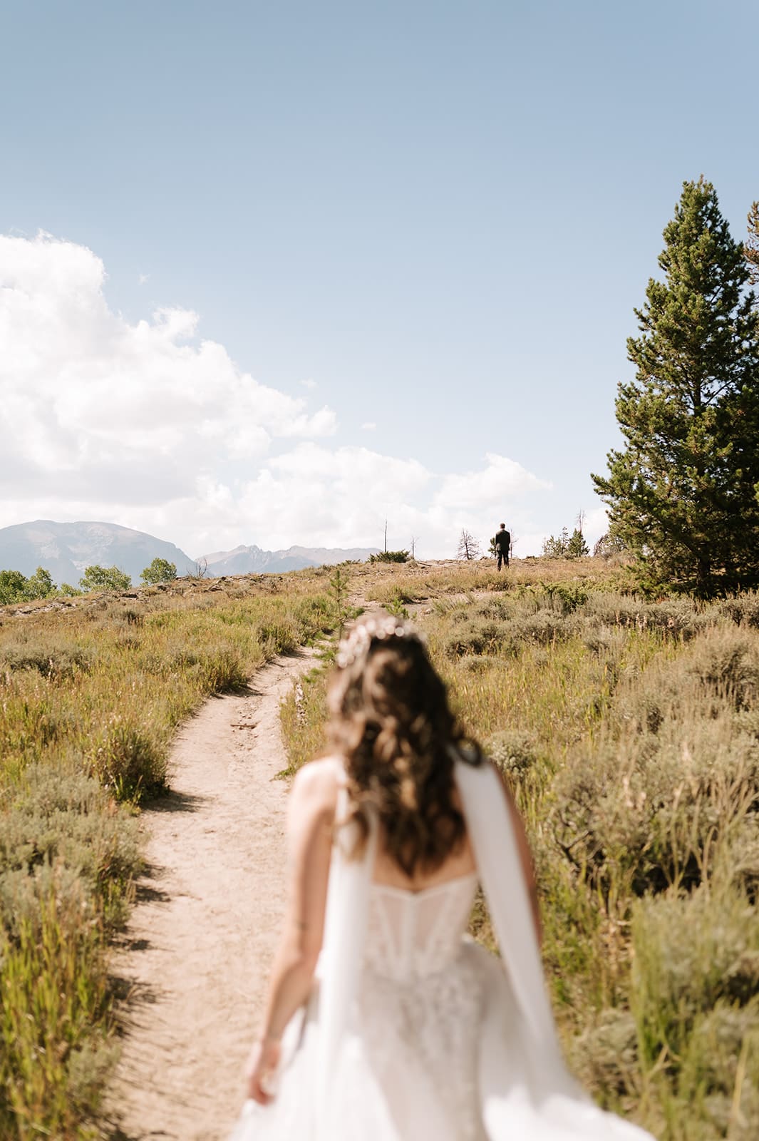 A woman in a white wedding dress walks along a dirt path through a grassy field under a clear sky.