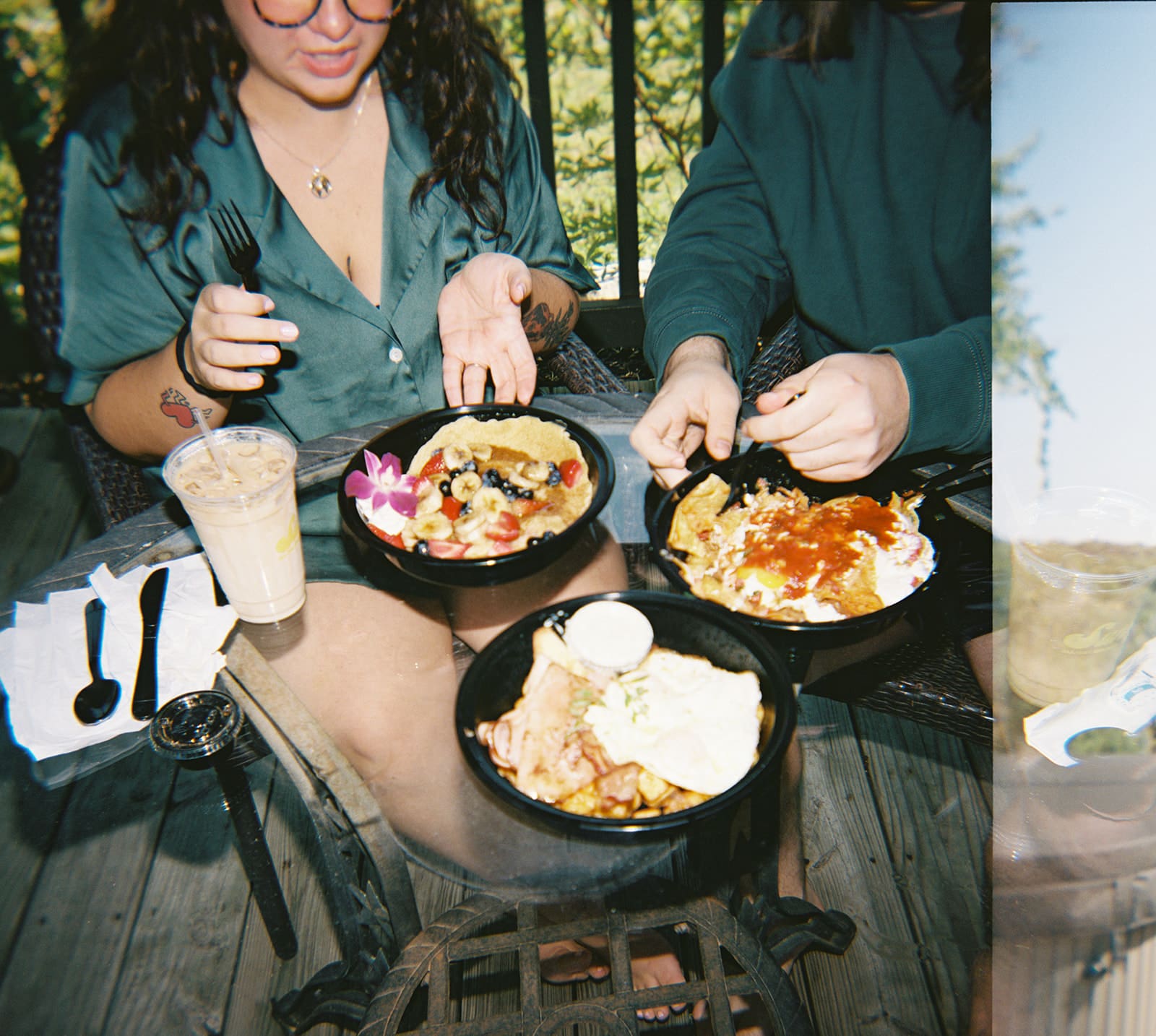 Two people sitting at an outdoor table with plates of food, including pancakes, hash browns, eggs, and a drink, preparing to eat.