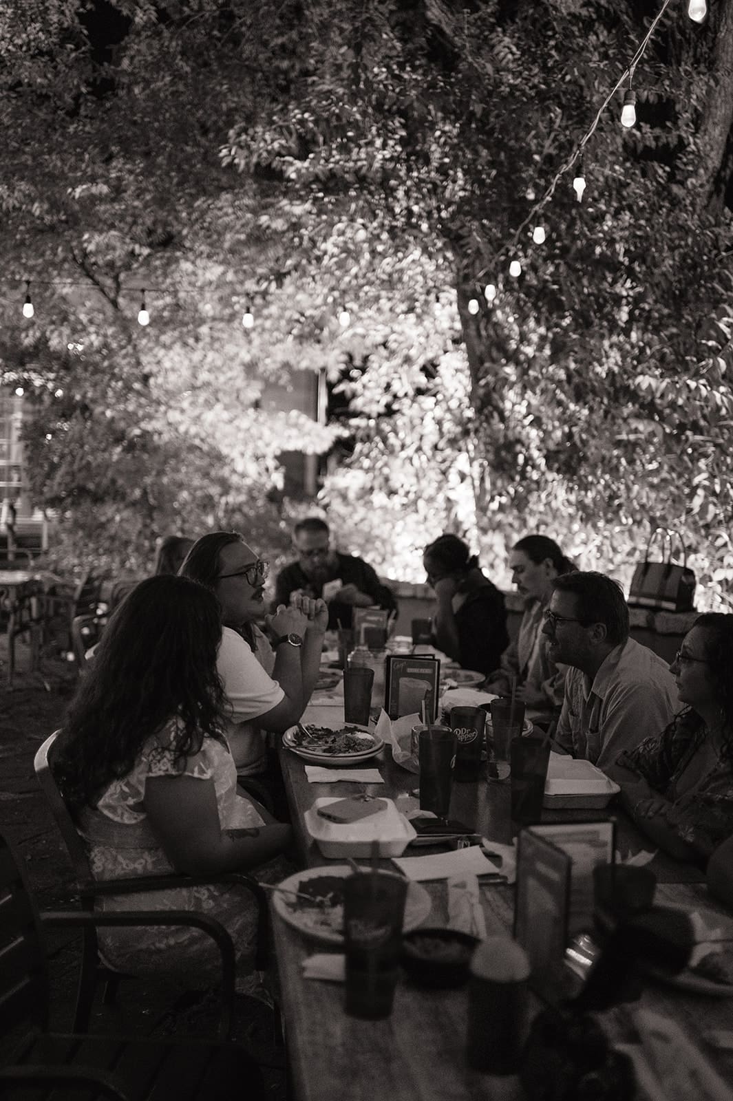 A group of twelve people sit around a long outdoor table at a restaurant, smiling for the camera, with menus and red cups in front of them.