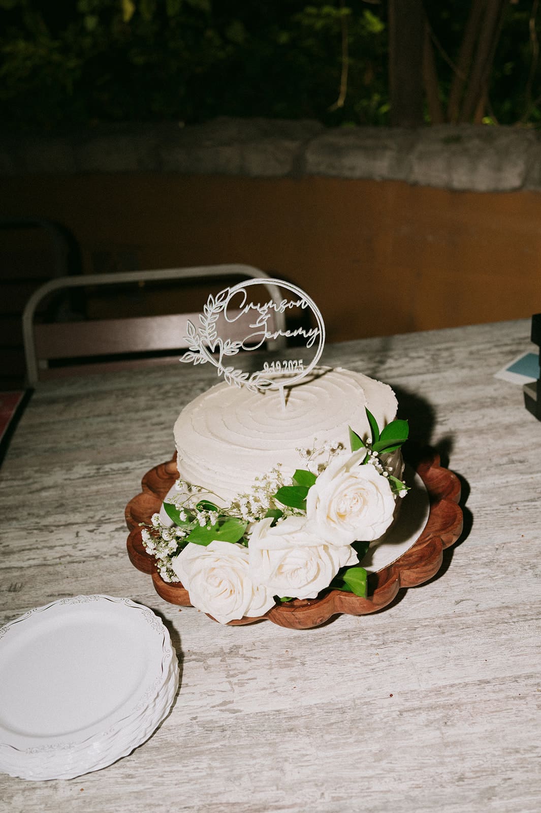 A white frosted cake decorated with white roses and greenery sits on a wooden cake stand near a stack of white plates on a table
