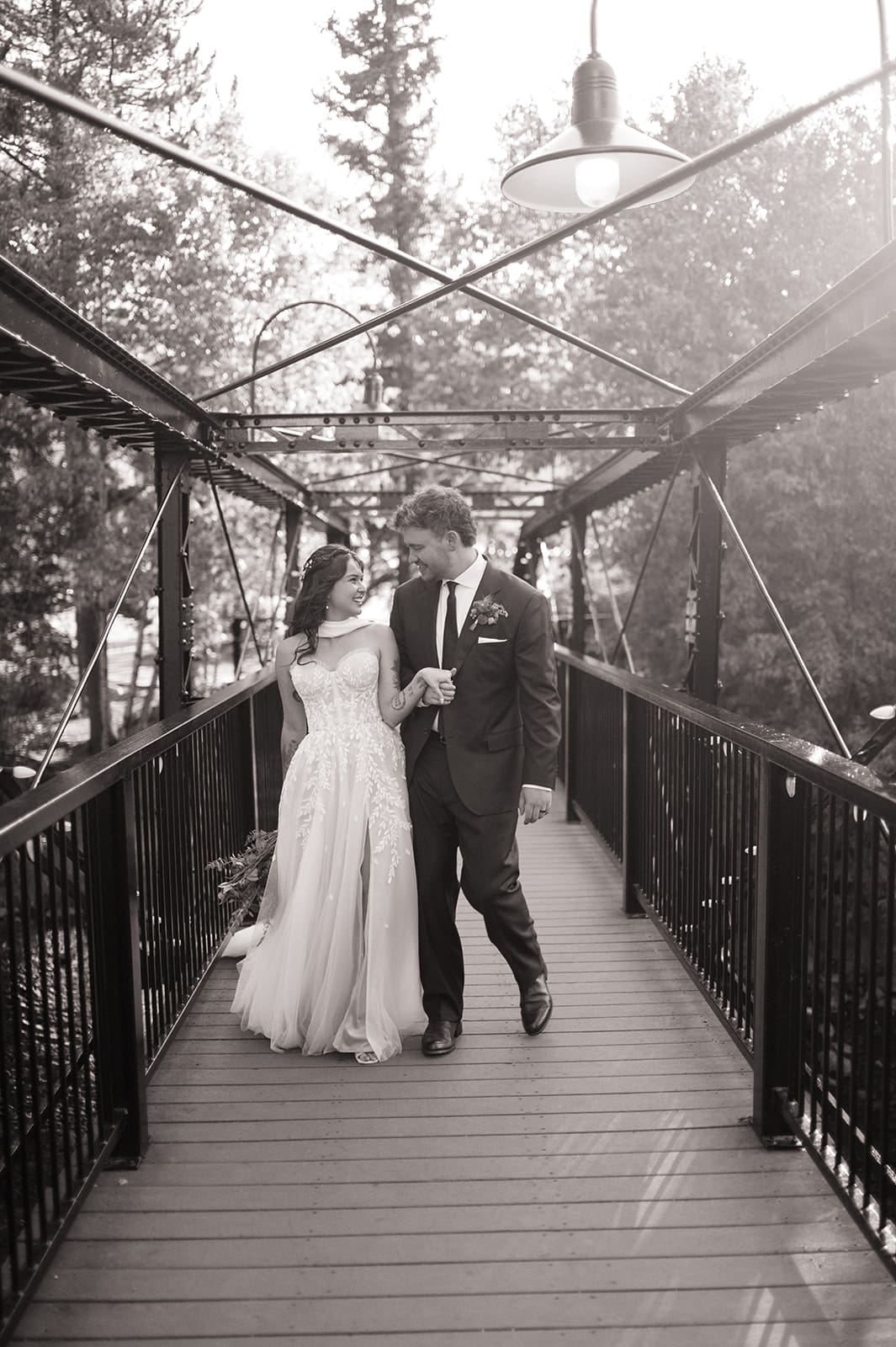 A bride in a white dress and a groom in a dark suit walk hand-in-hand on a metal bridge surrounded by trees.