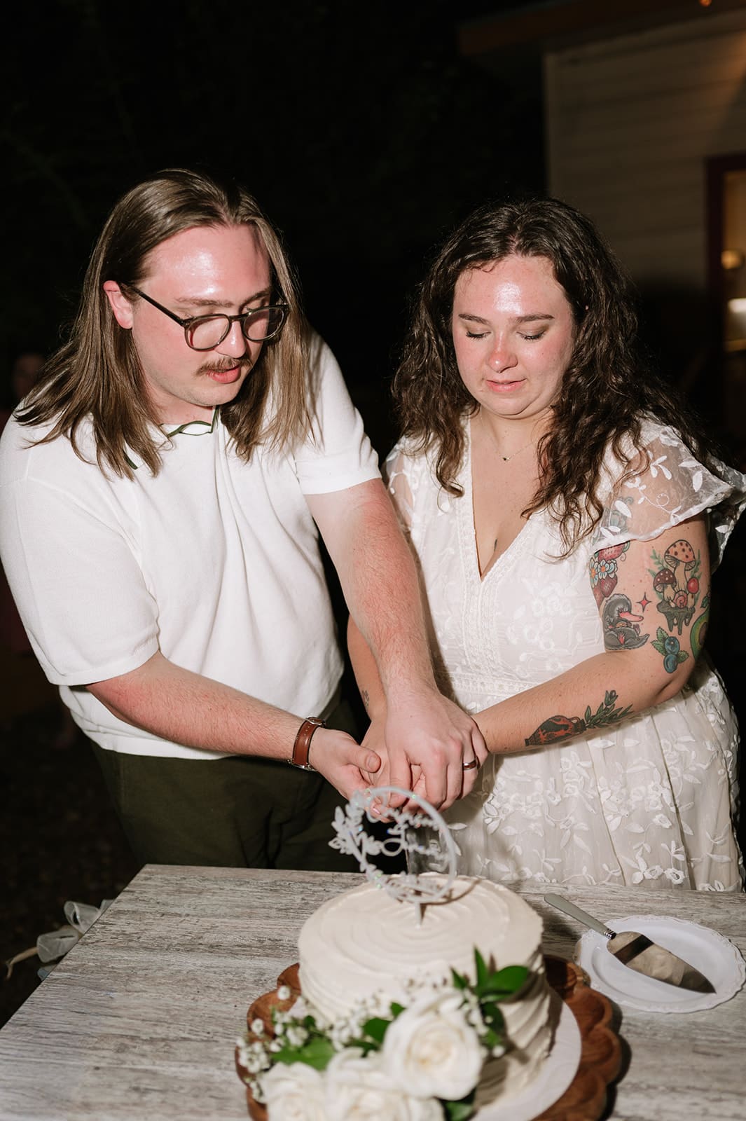 Two people stand outside at night, cutting into a wedding cake. One wears a white shirt, the other wears a white lace dress with visible tattoos.