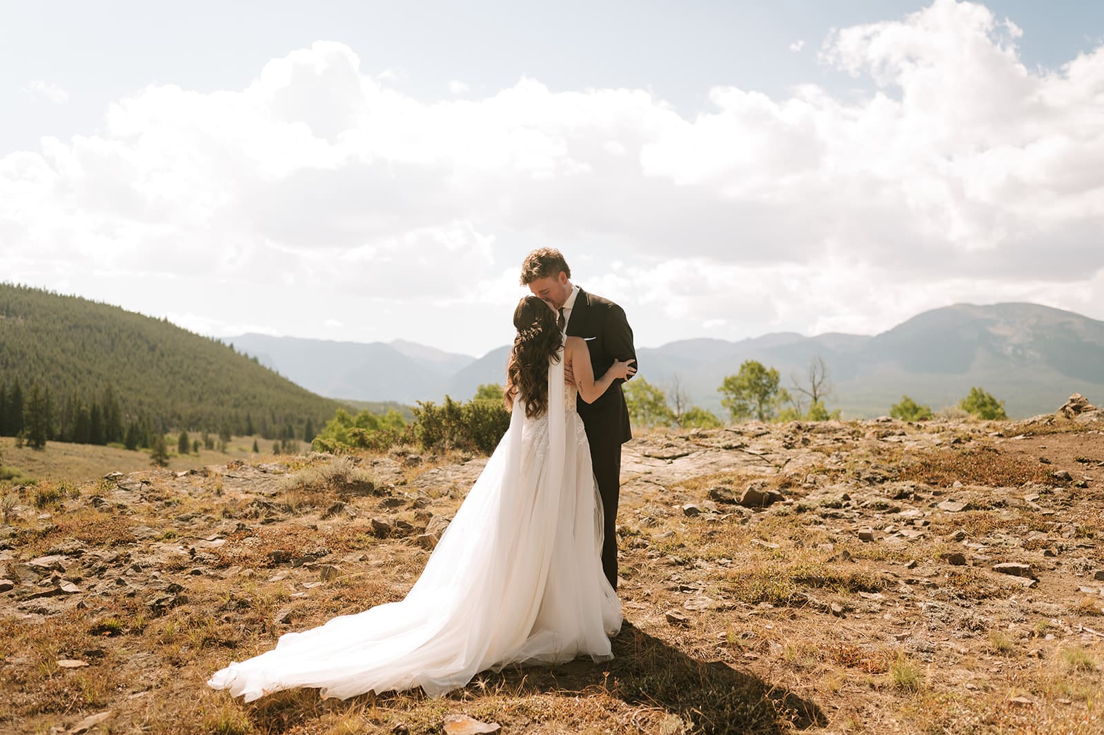 A bride and groom in wedding attire stand embracing on a rocky landscape with mountains and trees in the background under a partly cloudy sky for a Colorado wedding