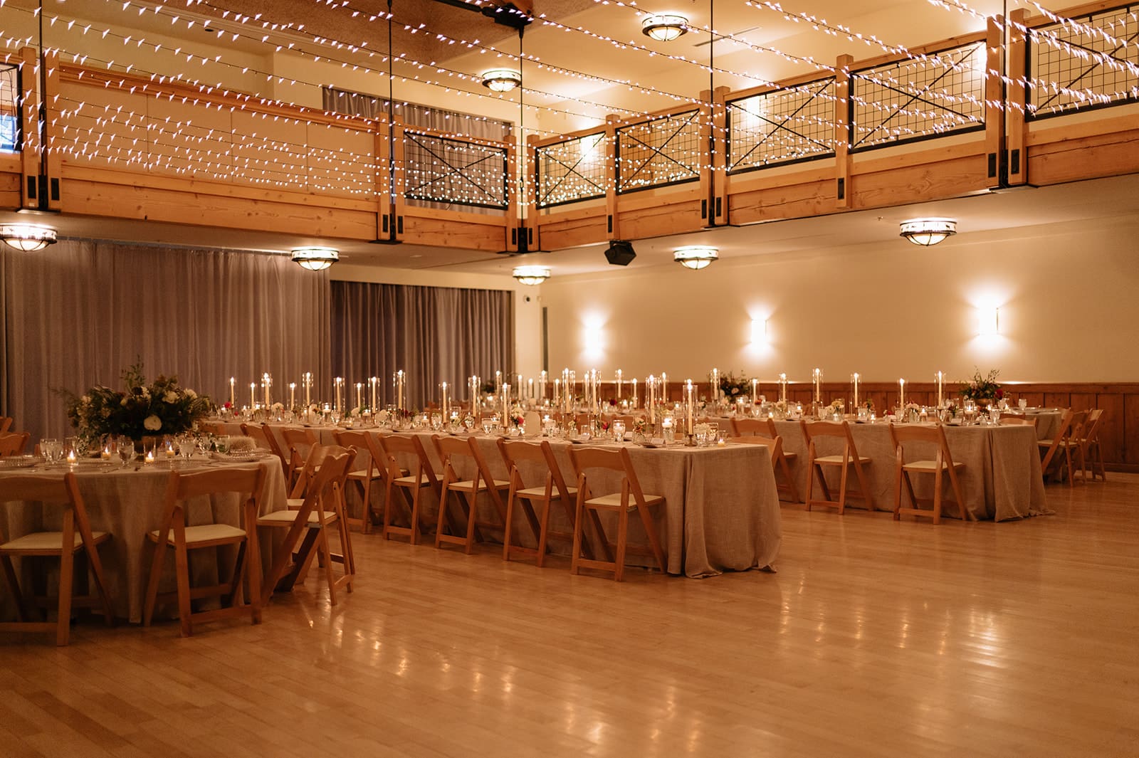 A long banquet table with wooden chairs, set with glassware, plates, and decorated with tall candles and small floral arrangements, is lit by candlelight in a dim room.