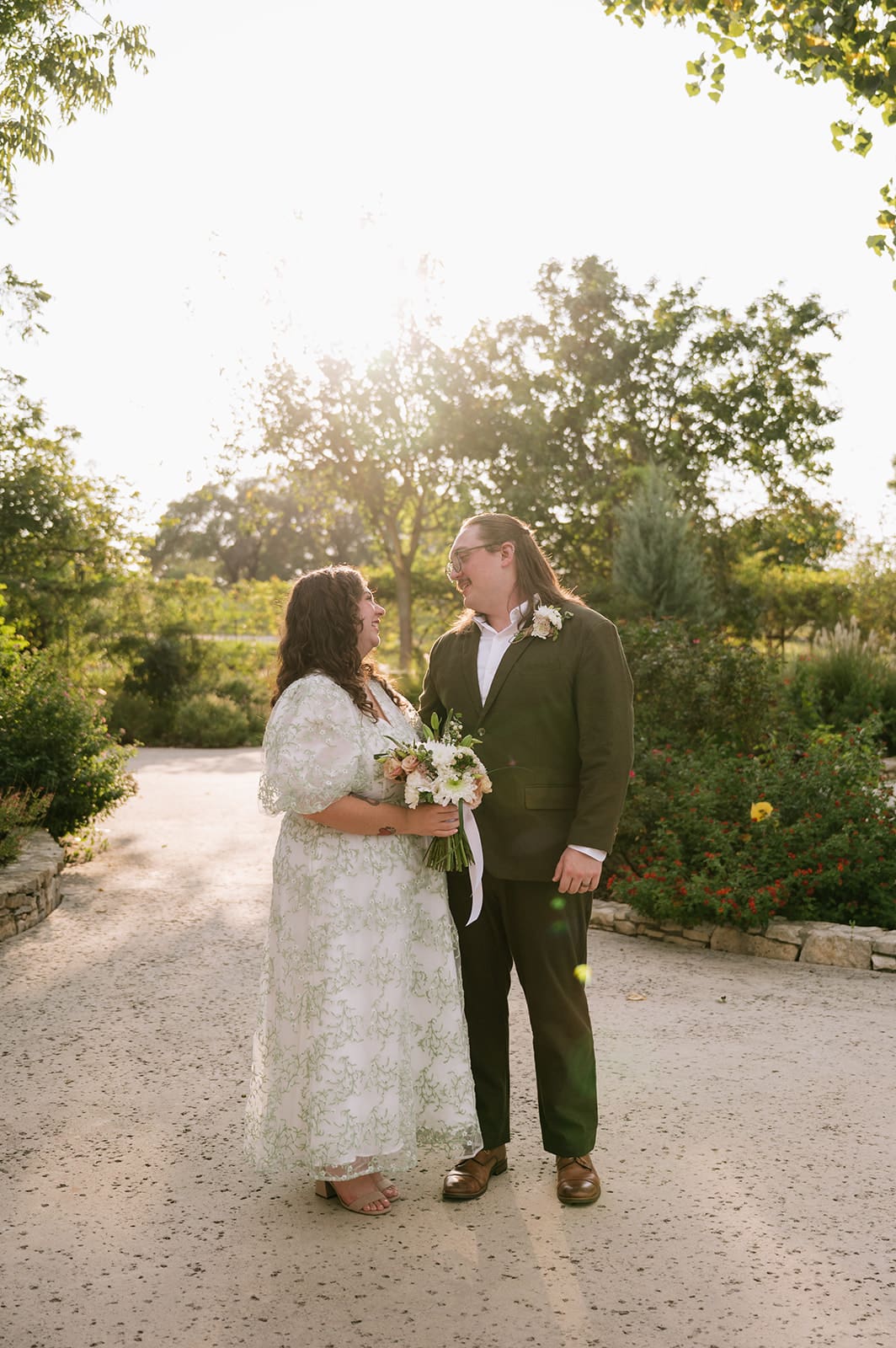 A couple dressed in wedding attire stands outdoors on a sunlit path, facing each other and holding a bouquet.