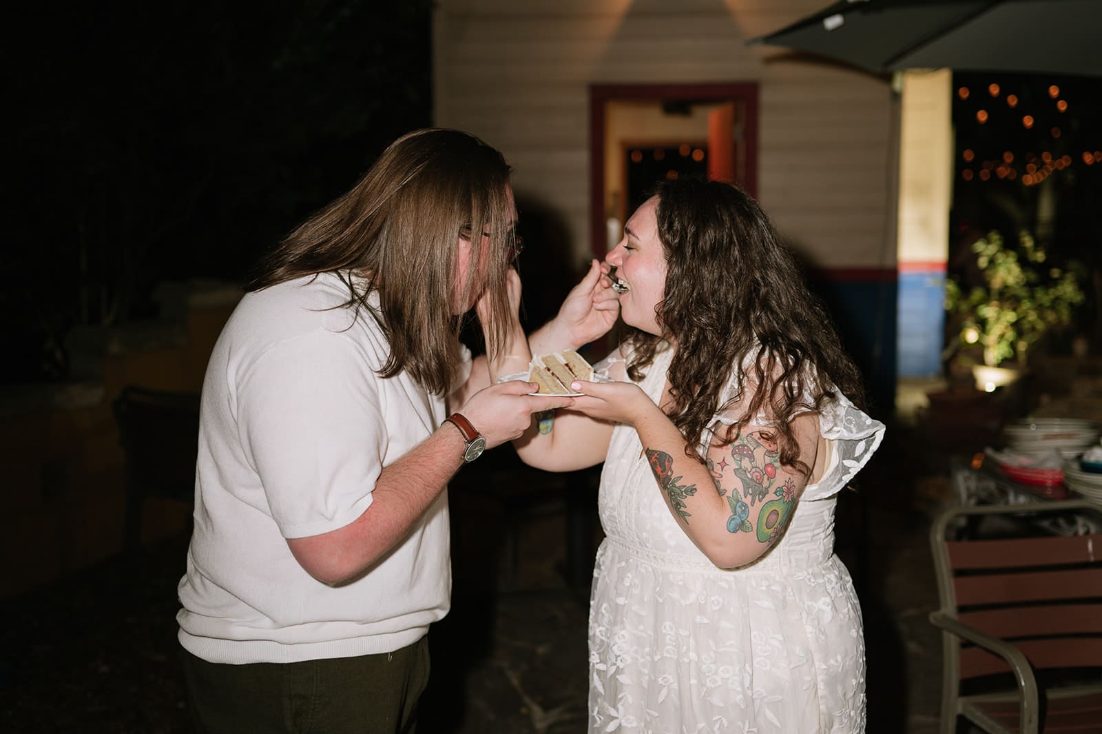 Two people stand outside at night, feeding each other cake and laughing. One wears a white shirt, the other wears a white lace dress with visible tattoos.