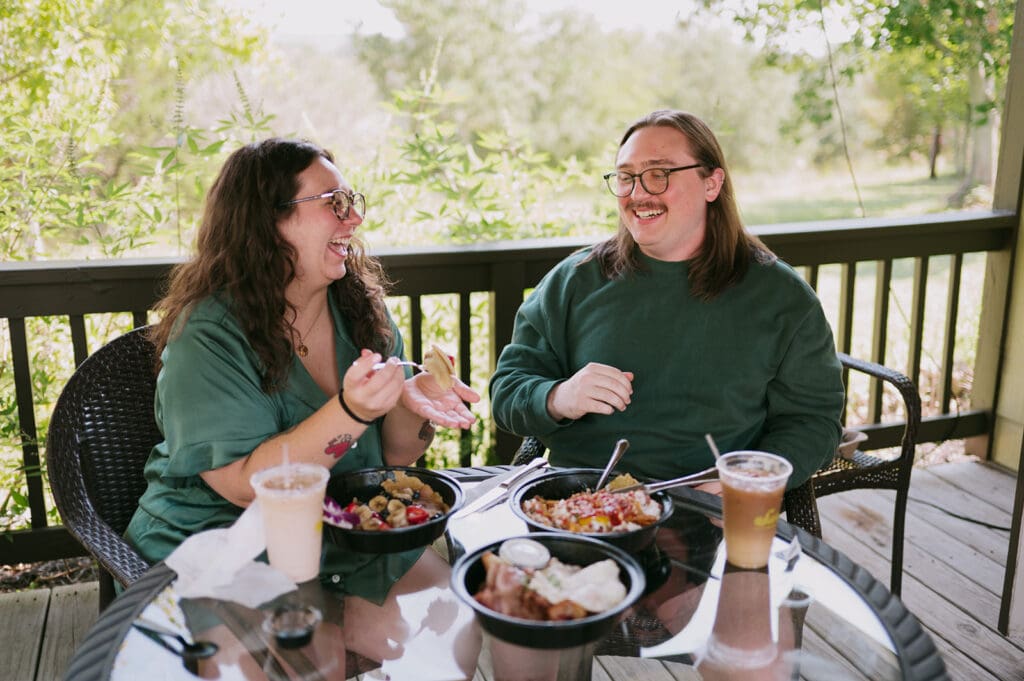 Two people sit at a round glass table on a wooden porch, eating and drinking together on a sunny day.