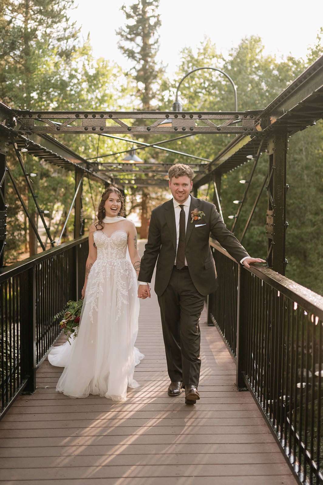 A bride in a white dress and a groom in a dark suit walk hand-in-hand on a metal bridge surrounded by trees.