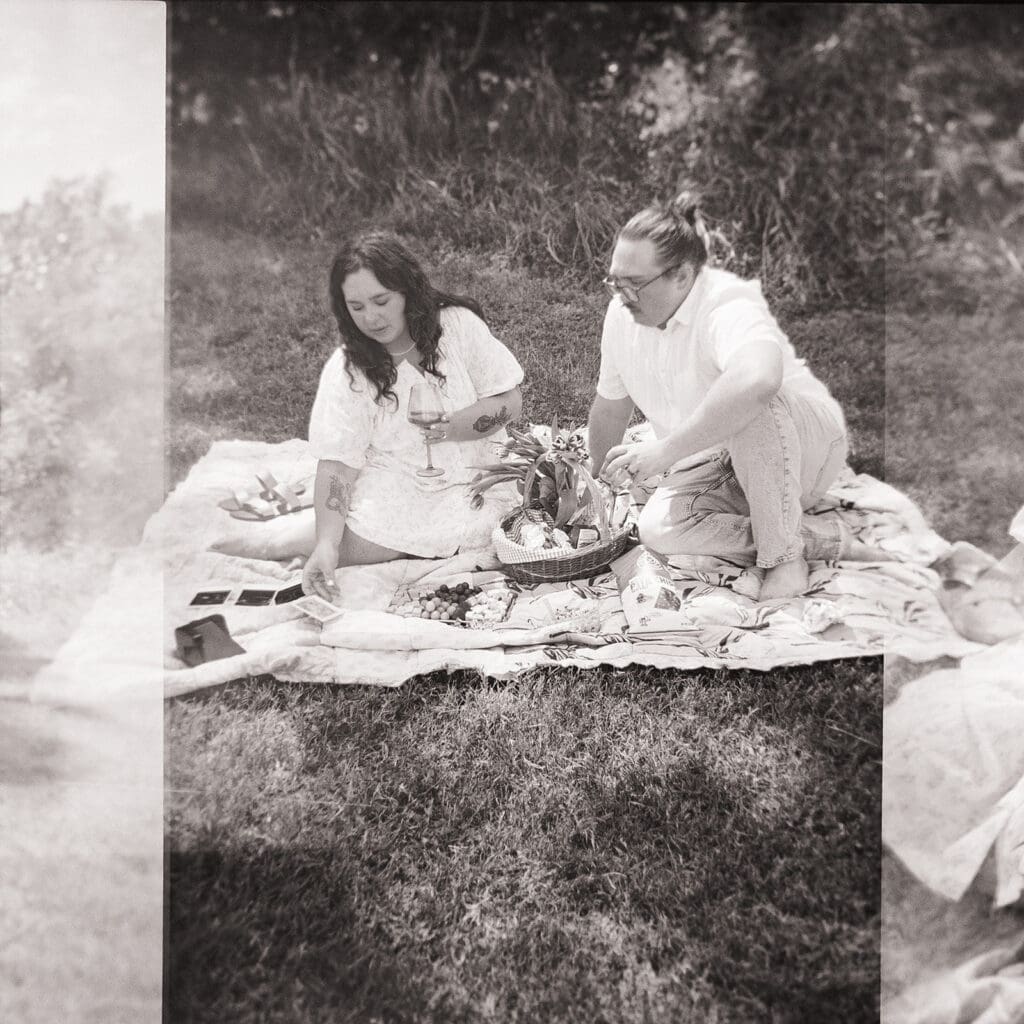 Two people sit on a blanket having a picnic outdoors, surrounded by greenery, with food and flowers arranged between them for a Texas Hill Country Elopement