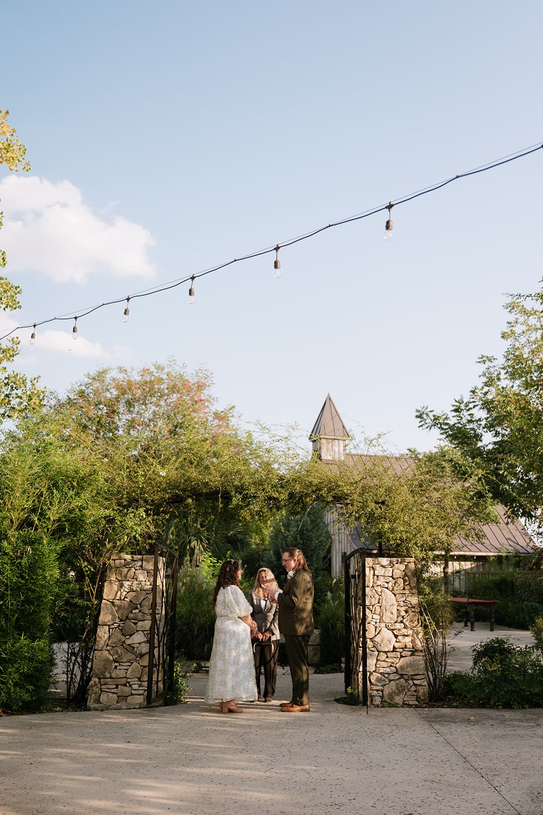 Two people stand facing each other outdoors under a leafy archway; one is holding a piece of paper and appears to be speaking for a texas hill country elopement