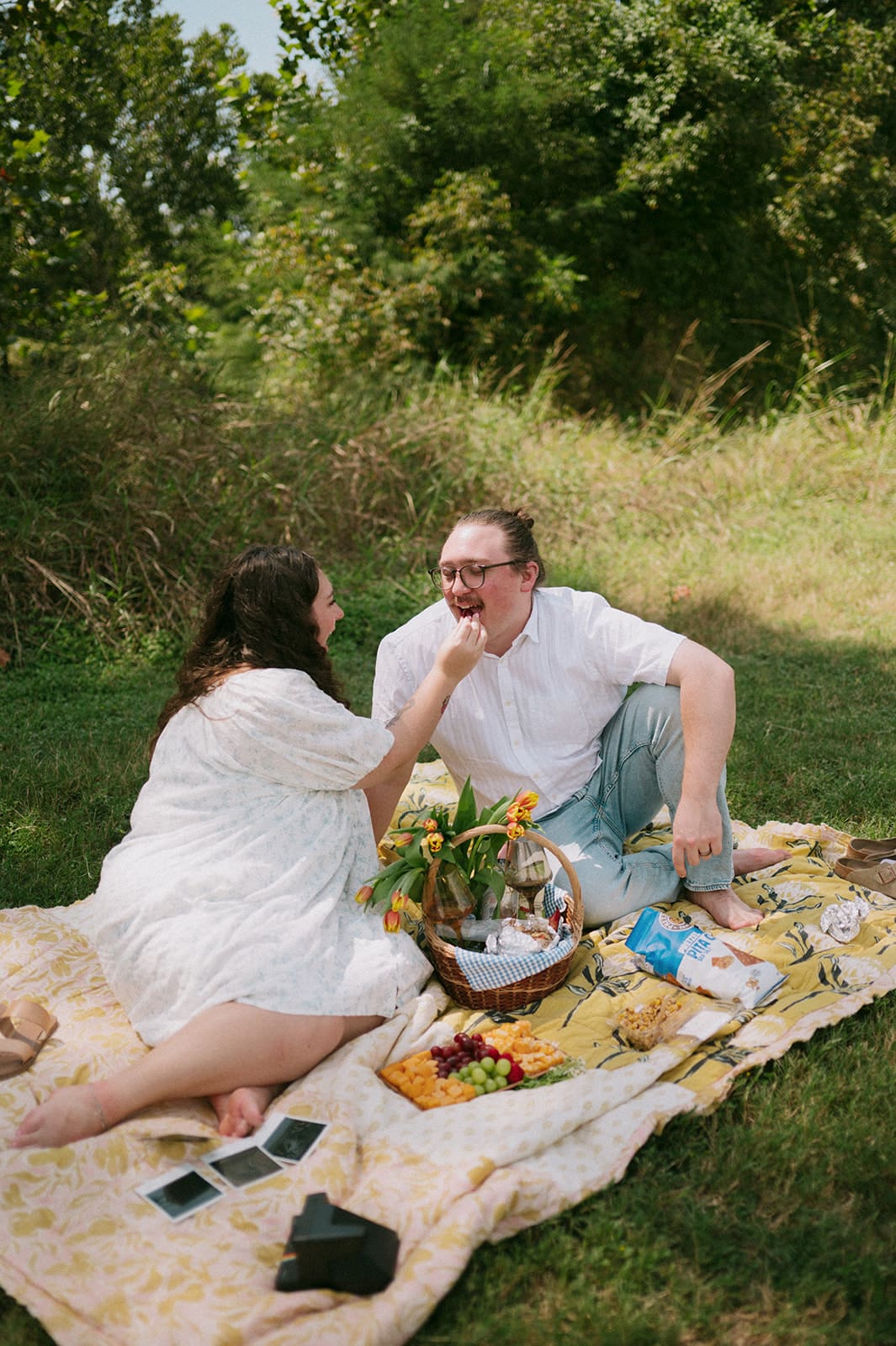 Two people sit on a blanket having a picnic outdoors, surrounded by greenery, with food and flowers arranged between them for a Texas Hill Country Elopement
