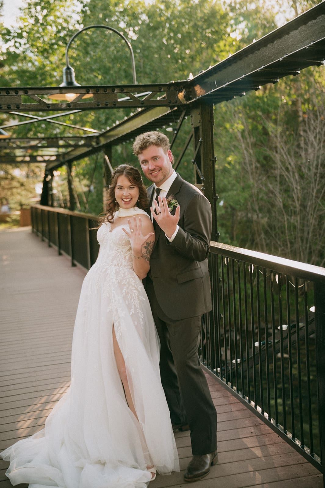 A bride in a white dress and a groom in a dark suit walk hand-in-hand on a metal bridge surrounded by trees.