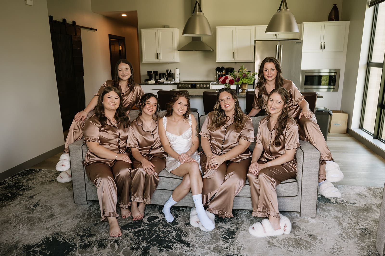 Seven women sit together on a couch in a modern living room; one is wearing white, and the others are in matching brown satin pajamas.