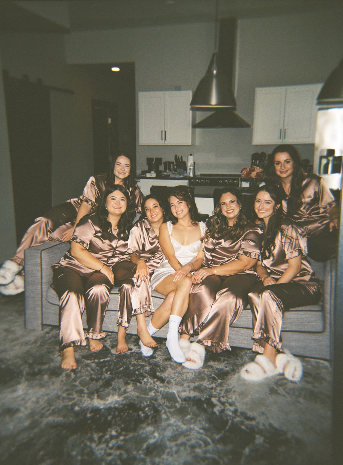 Seven women sit together on a couch in a modern living room; one is wearing white, and the others are in matching brown satin pajamas.