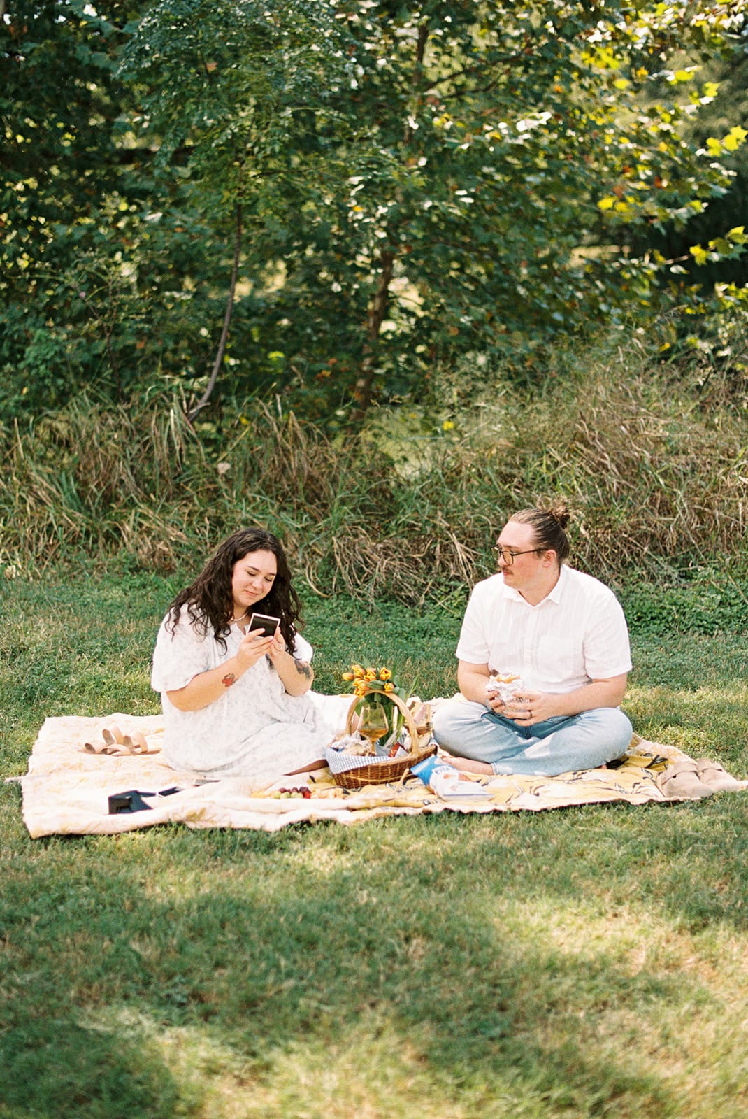 Two people sit on a blanket having a picnic outdoors, surrounded by greenery, with food and flowers arranged between them for a Texas Hill Country Elopement