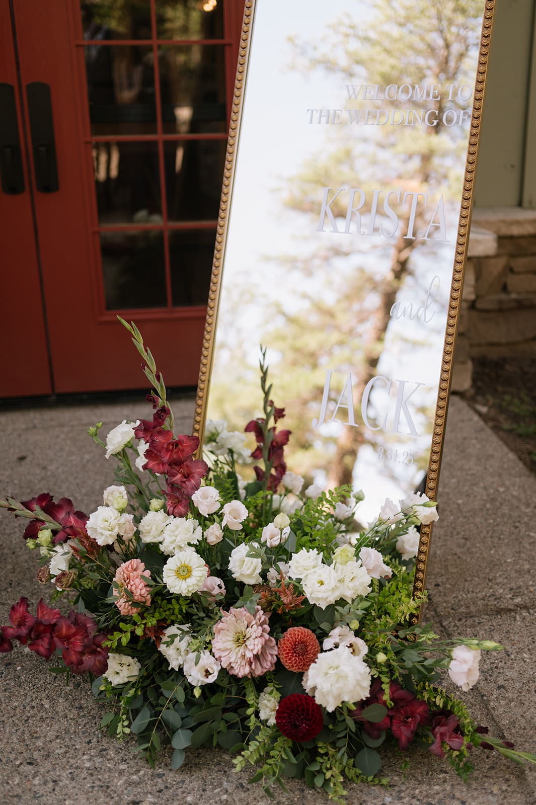 A large gold-framed mirror with white writing stands outdoors beside a building, with a floral arrangement of mixed flowers at its base.