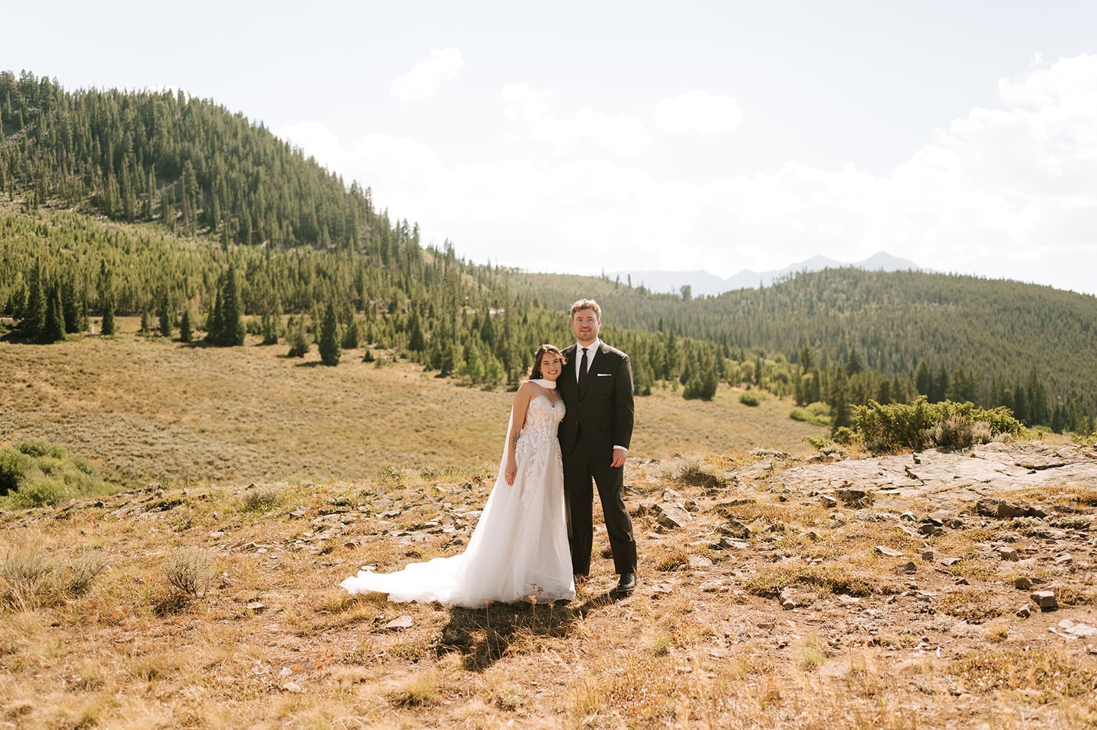 A bride and groom in wedding attire stand embracing on a rocky landscape with mountains and trees in the background under a partly cloudy sky for a Colorado wedding