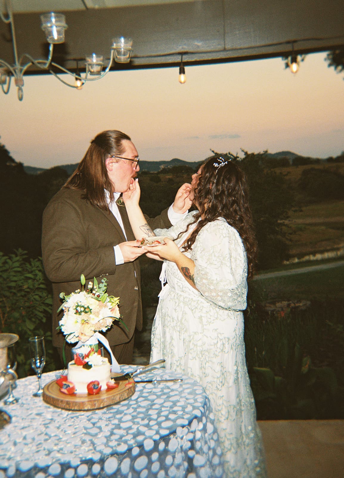 Two people, dressed formally, cut a small decorated cake together on a patterned tablecloth, with a bouquet of flowers nearby.