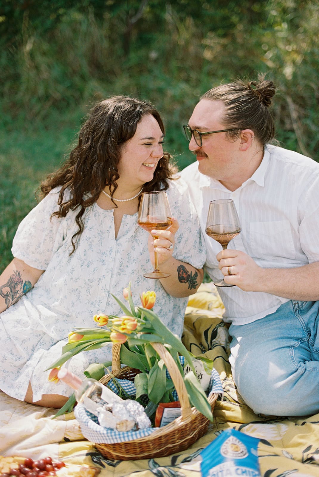 Two people sit on a blanket having a picnic outdoors, surrounded by greenery, with food and flowers arranged between them for a Texas Hill Country Elopement