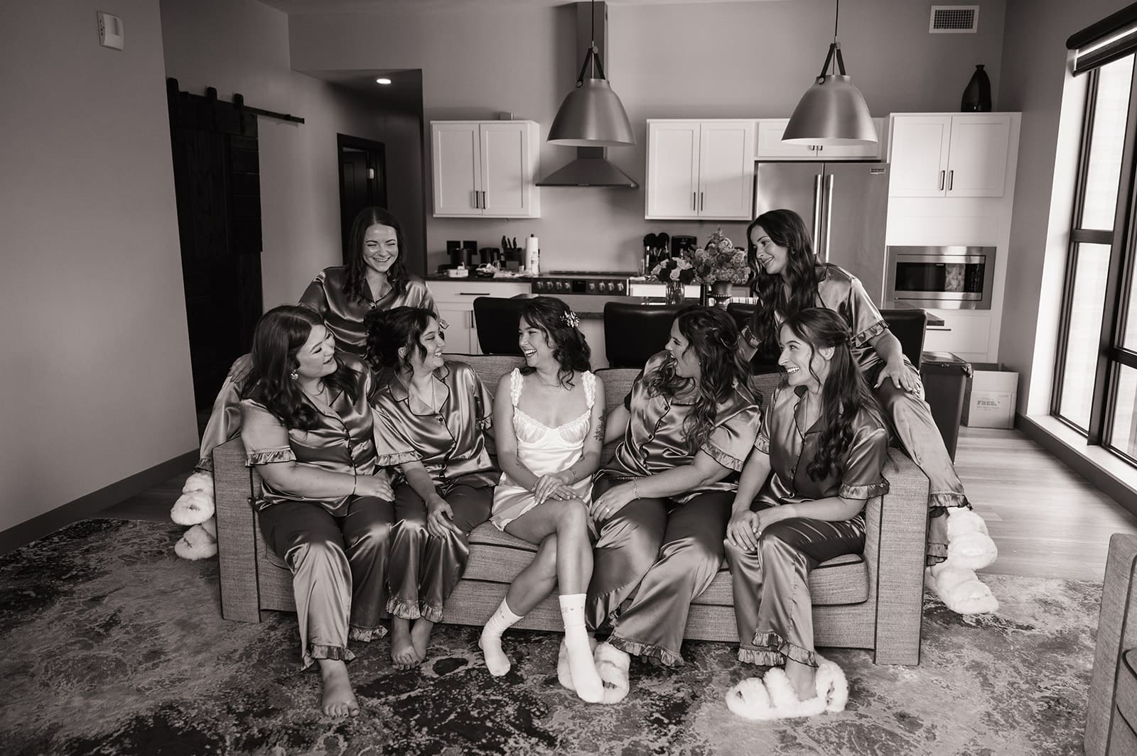 Seven women sit together on a couch in a modern living room; one is wearing white, and the others are in matching brown satin pajamas.