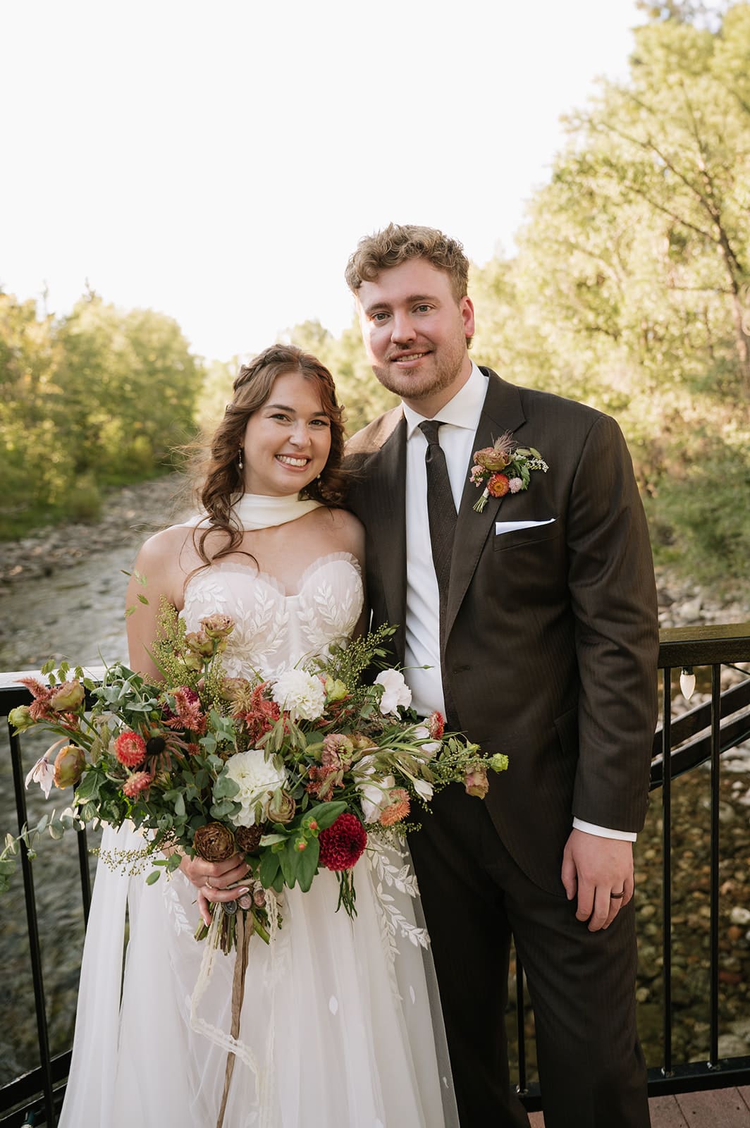 A bride in a white dress and a groom in a dark suit walk hand-in-hand on a metal bridge surrounded by trees.