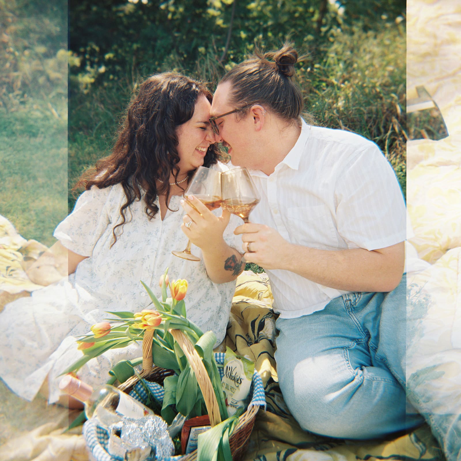 Two people sit on a blanket having a picnic outdoors, surrounded by greenery, with food and flowers arranged between them for a Texas Hill Country Elopement