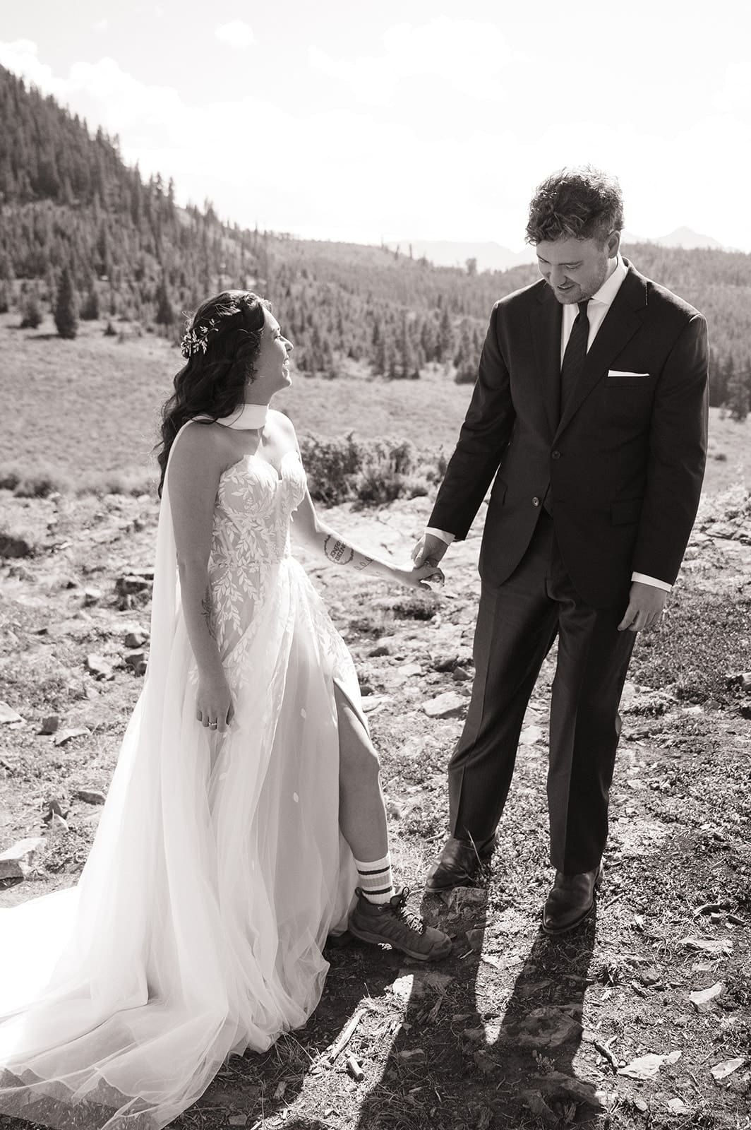 A bride and groom in wedding attire stand embracing on a rocky landscape with mountains and trees in the background under a partly cloudy sky for a Colorado wedding