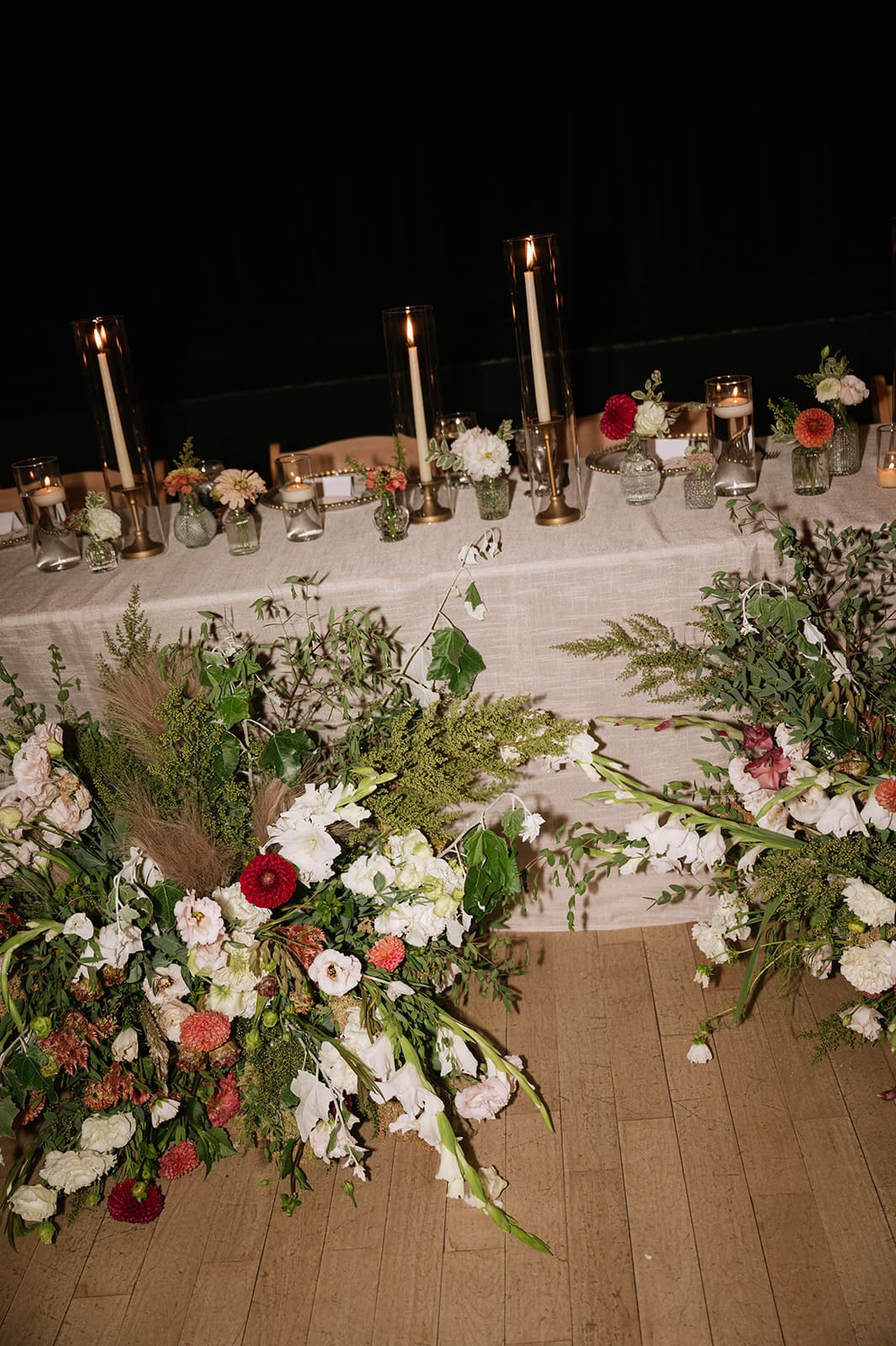 A long banquet table with wooden chairs, set with glassware, plates, and decorated with tall candles and small floral arrangements, is lit by candlelight in a dim room.