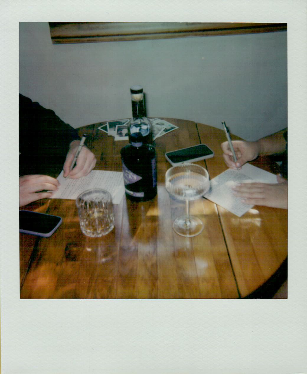 Two people sit at a wooden table with drinks, paper, and pens in hand. There are two phones and a bottle on the table.