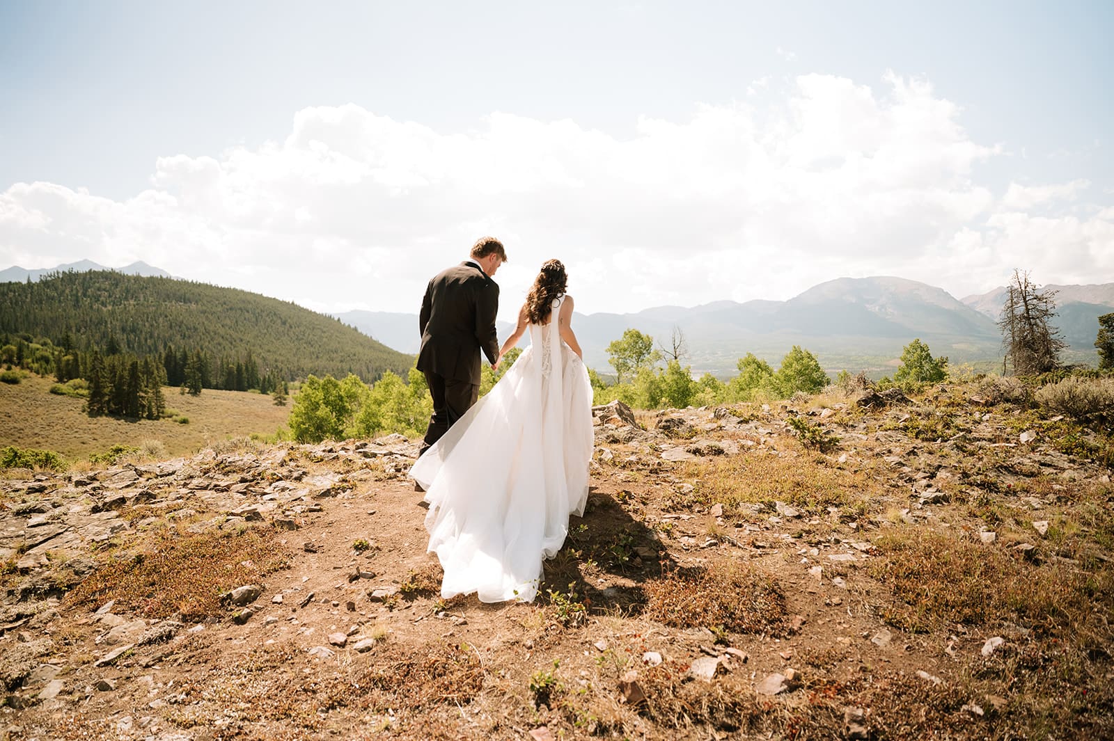 A bride and groom in wedding attire stand embracing on a rocky landscape with mountains and trees in the background under a partly cloudy sky for a Colorado wedding