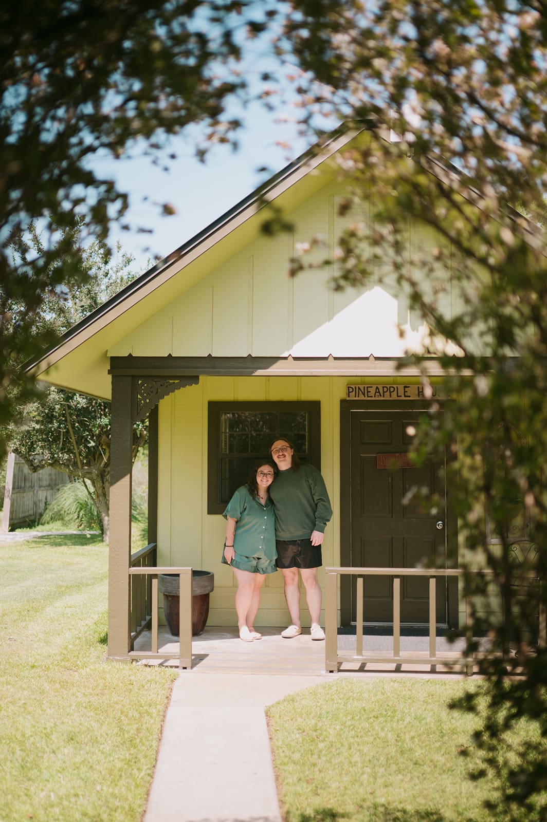 Two people stand together on the front porch of a small, light-colored house surrounded by greenery on a sunny day.