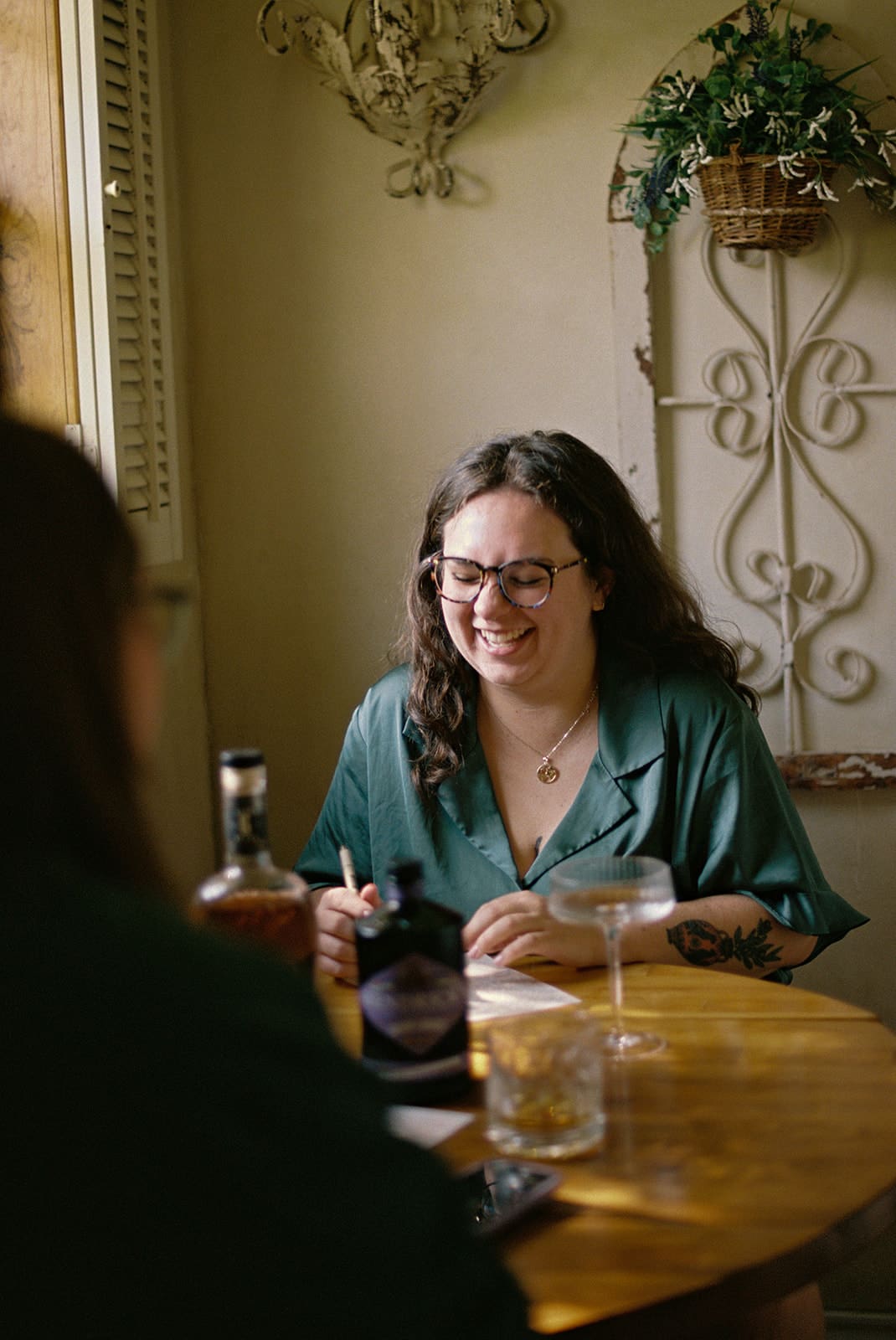 Two people sit at a small wooden table with drinks and bottles, engaged in conversation by a window in a softly lit room.