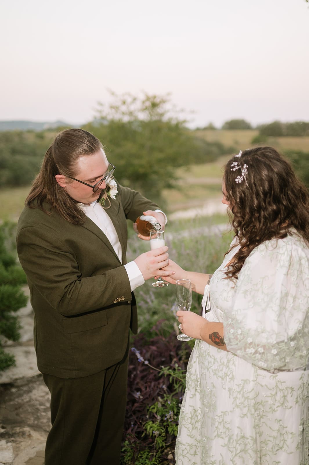 Two people, dressed formally, cut a small decorated cake together on a patterned tablecloth, with a bouquet of flowers nearby.