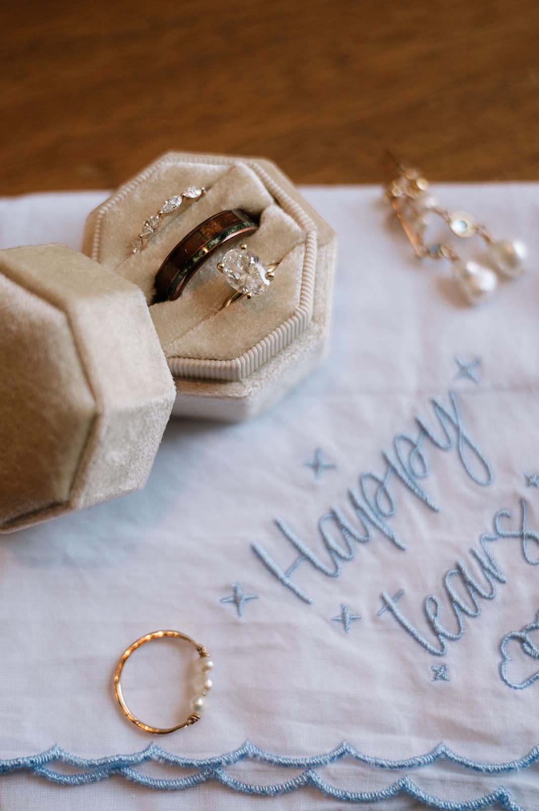A pair of beige high-heeled sandals, a blue embroidered handkerchief with wedding rings, a jewelry box, and a bouquet of flowers arranged on a wooden surface.