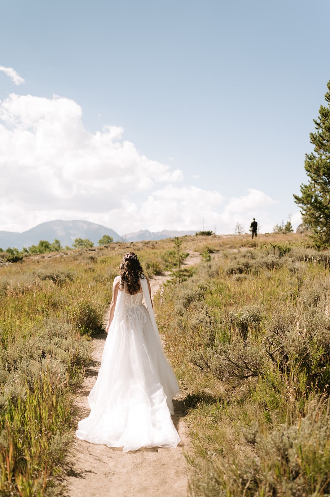 A woman in a white wedding dress walks along a dirt path through a grassy field under a clear sky.