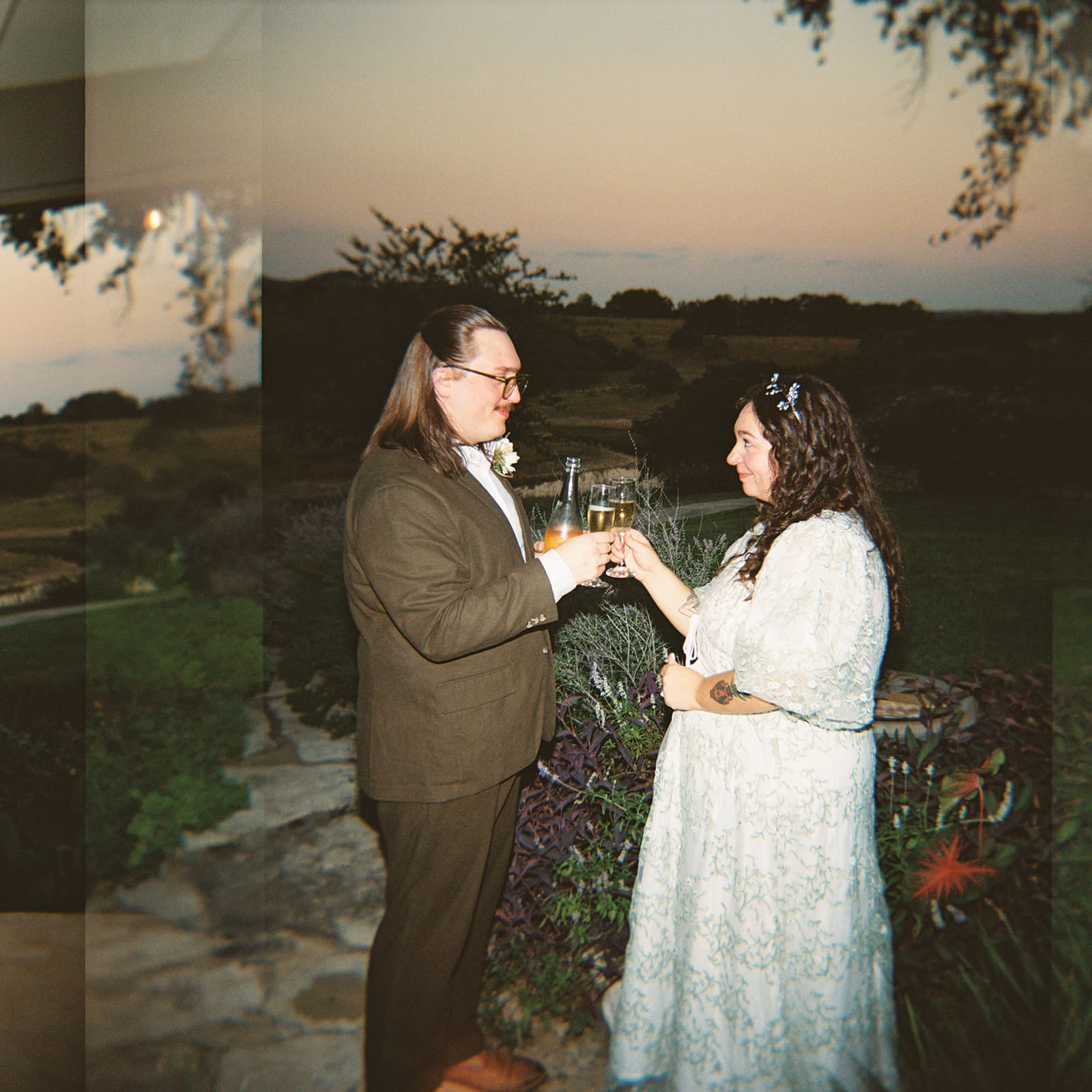 Two people dressed formally clink glasses and smile at each other outdoors at sunset, surrounded by greenery and landscape views for a texas hill country elopement
