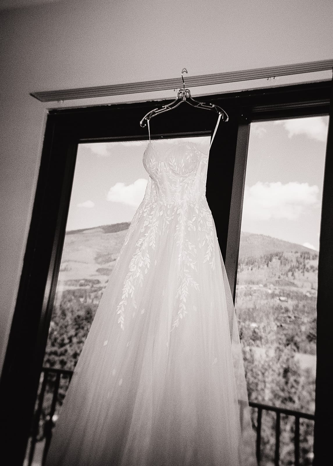 A white strapless wedding dress hangs on a hanger by a window with a blurred outdoor landscape in the background.