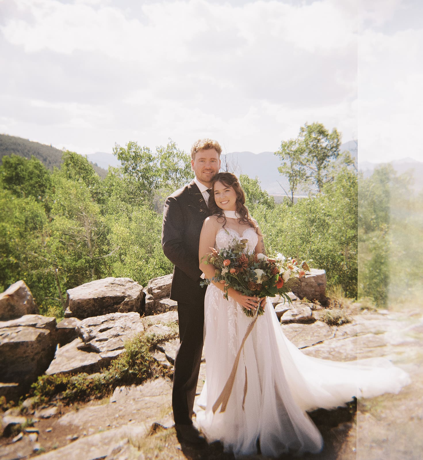 A bride and groom in wedding attire stand embracing on a rocky landscape with mountains and trees in the background under a partly cloudy sky for a Colorado wedding