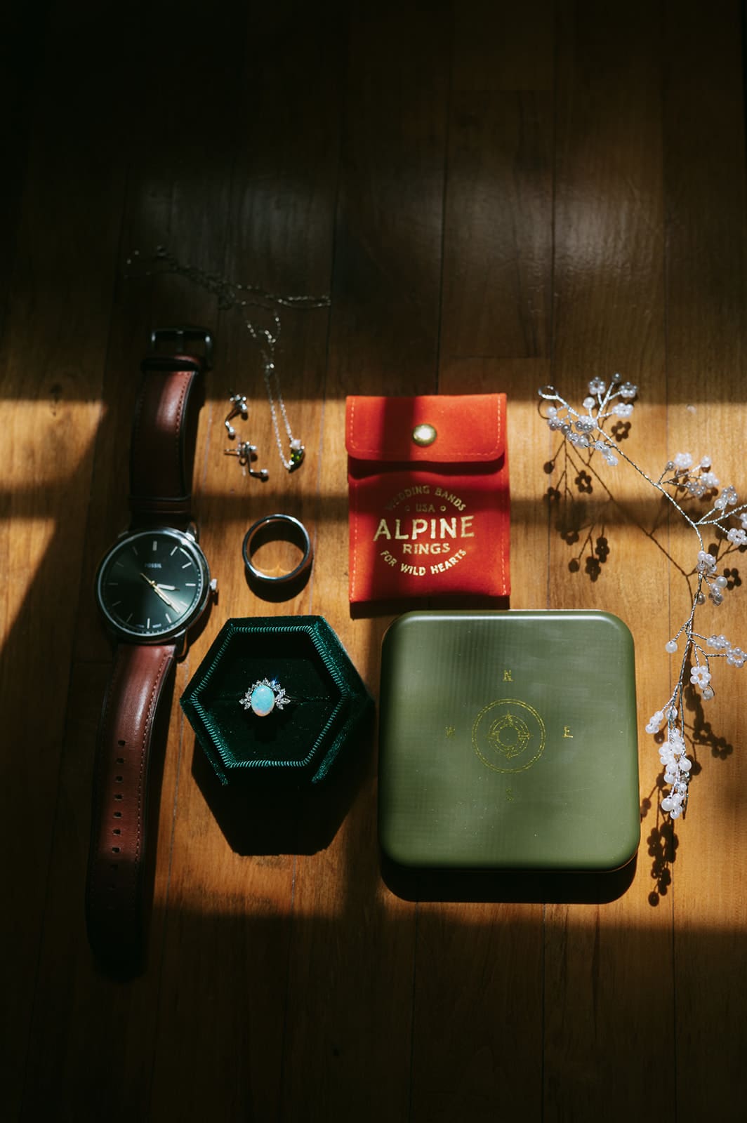 A wristwatch, two rings, a green jewelry box, a red pouch labeled "ALPINE," and small decorative branches are arranged on a wooden surface with sunlight.