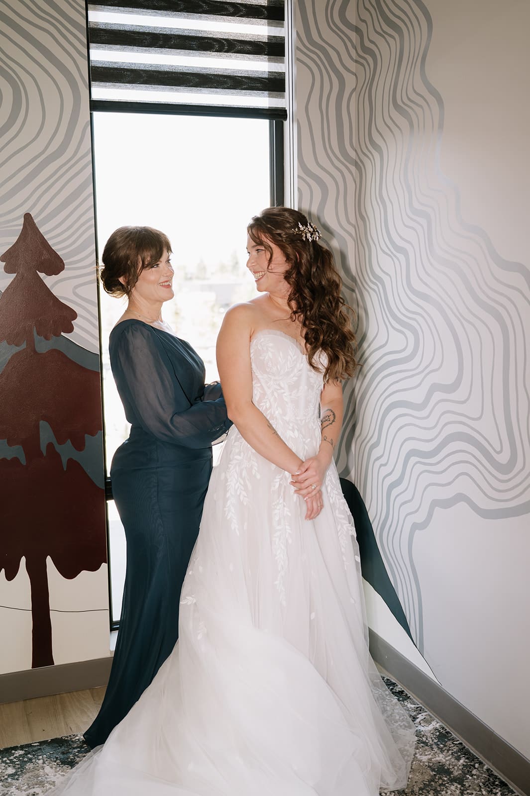 Two women stand indoors by a window; one wears a dark dress and the other wears a white wedding gown. They face each other and smile.