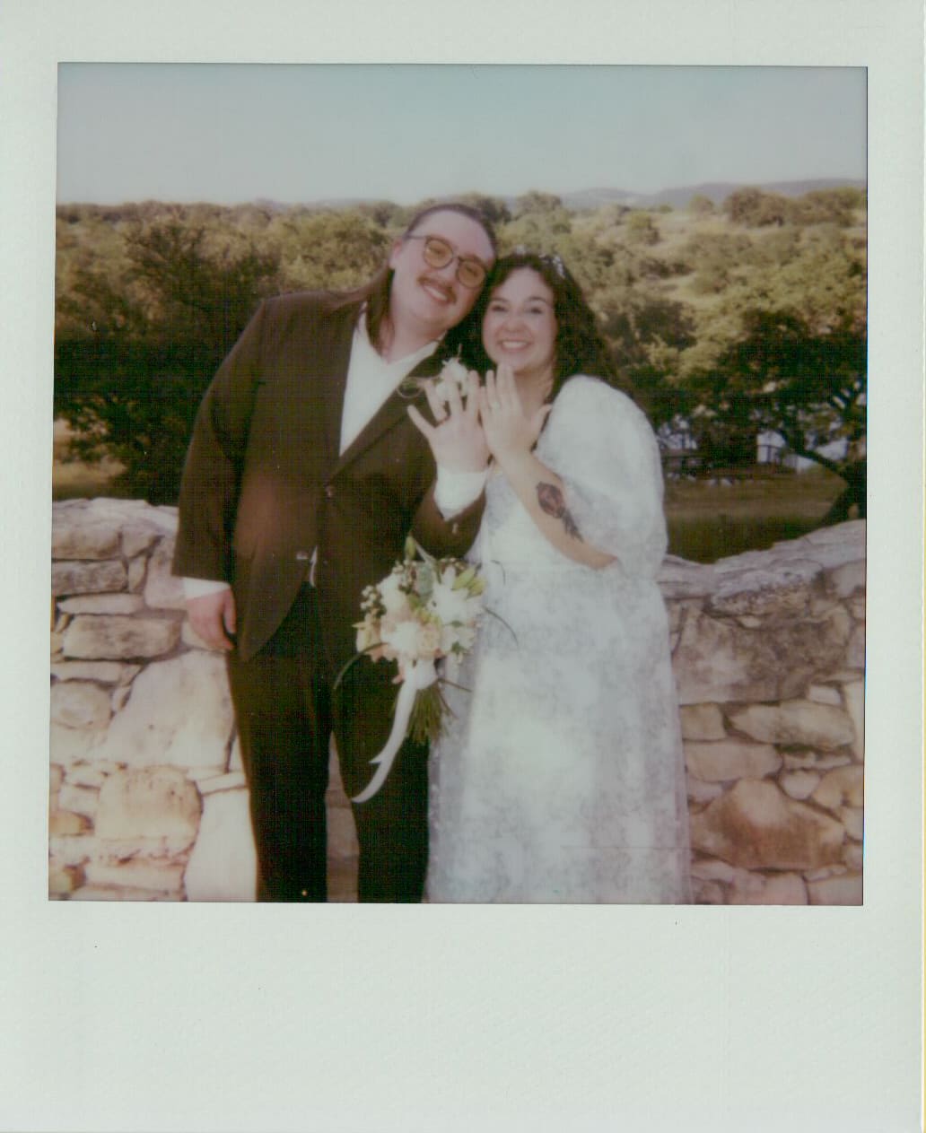 A couple poses outdoors by a stone wall, smiling and showing their rings. One person holds a bouquet and both wear formal attire. Trees are visible in the background.