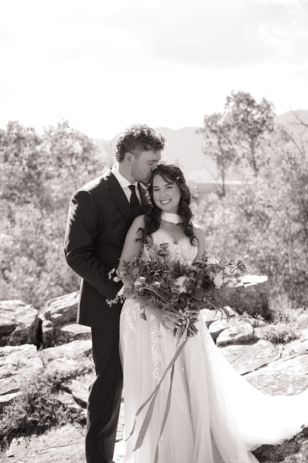 A bride and groom in wedding attire stand embracing on a rocky landscape with mountains and trees in the background under a partly cloudy sky for a Colorado wedding