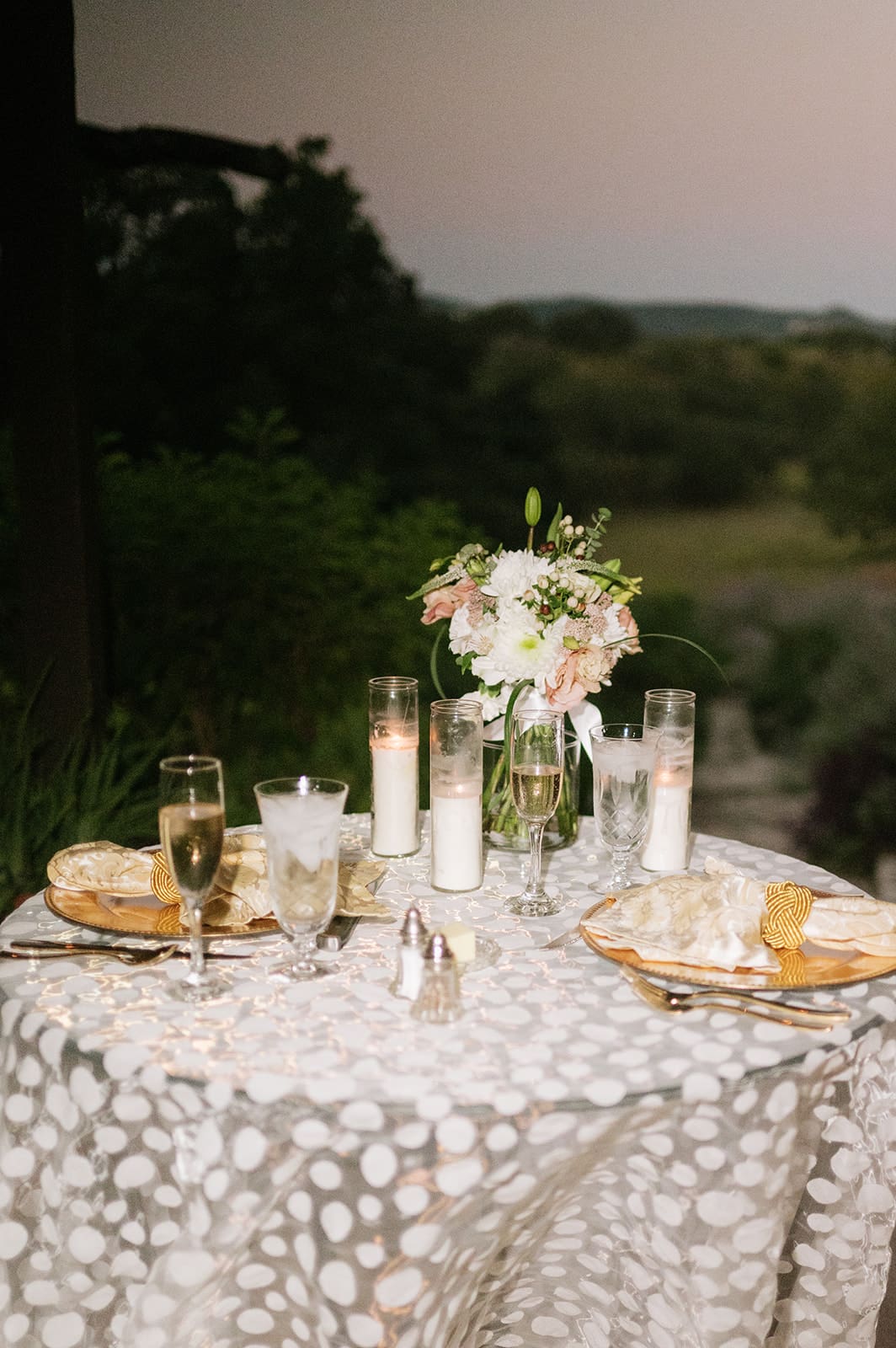 Two people, dressed formally, cut a small decorated cake together on a patterned tablecloth, with a bouquet of flowers nearby.