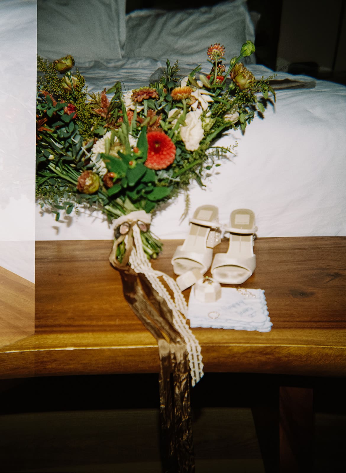 A pair of beige high-heeled sandals, a blue embroidered handkerchief with wedding rings, a jewelry box, and a bouquet of flowers arranged on a wooden surface.