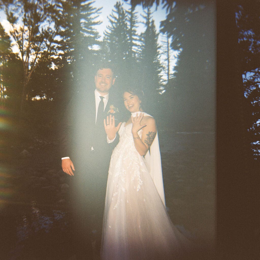 A couple dressed in formal wedding attire stands outdoors, with the woman showing her ring. Tall trees are visible in the background. The image is slightly faded and has a light flare effect.