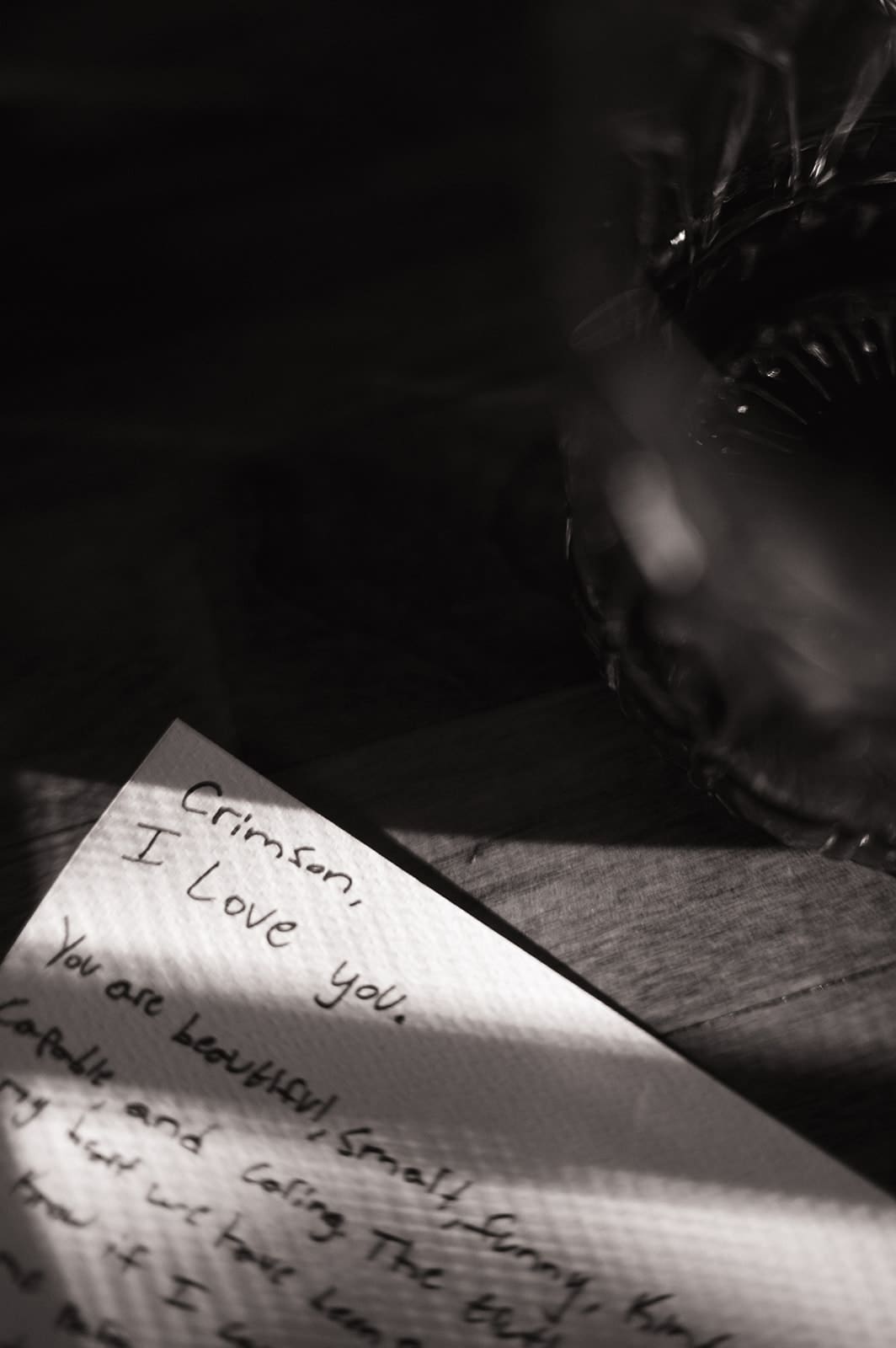 A handwritten note on textured paper reads "Crimson, I love you" with more cursive text below, partially obscured by shadows and a glass object in the upper right.