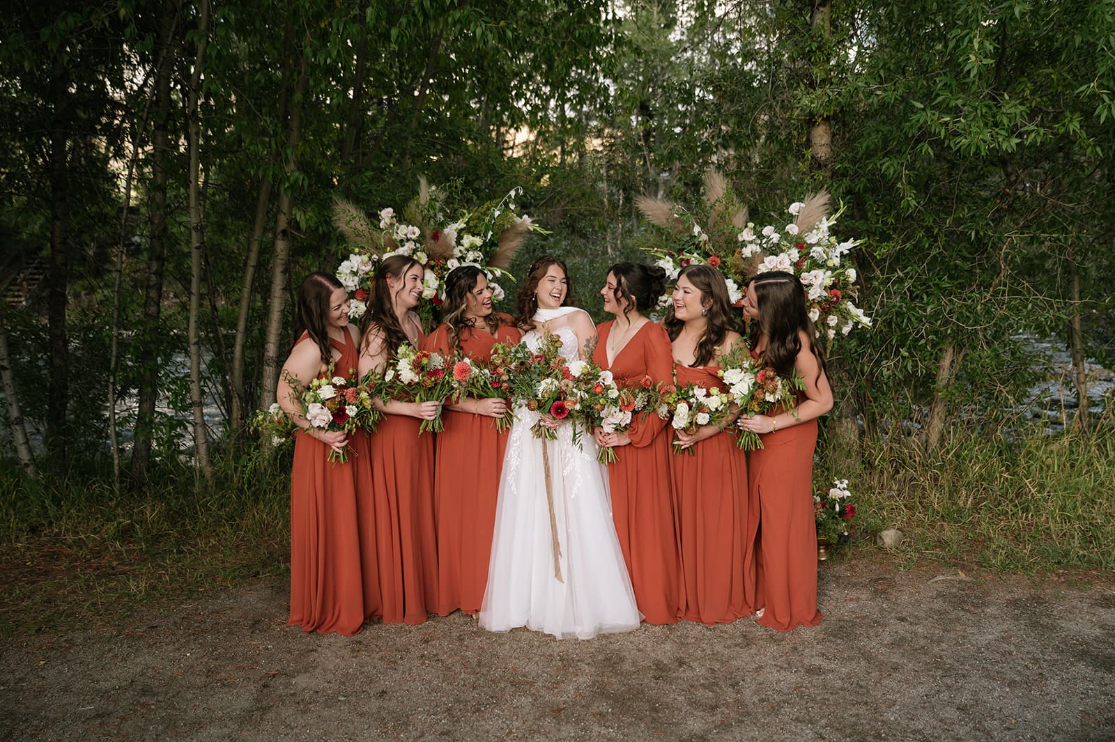 Five women stand together outdoors, one in a white wedding dress and four in matching rust-colored dresses, all holding bouquets of flowers and smiling.