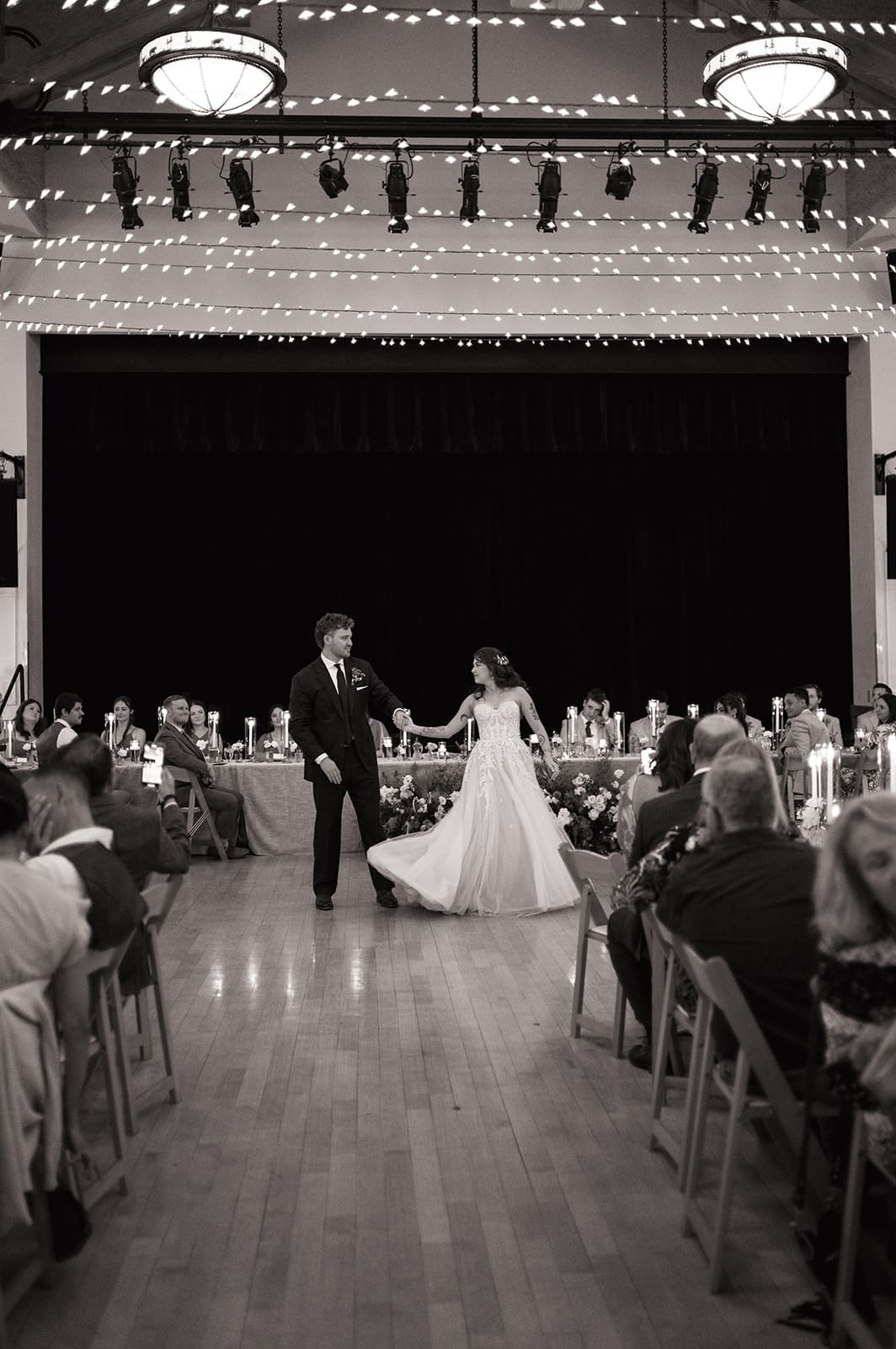 A bride and groom dance in the center of a large hall with guests seated at tables, string lights overhead, and a stage in the background.