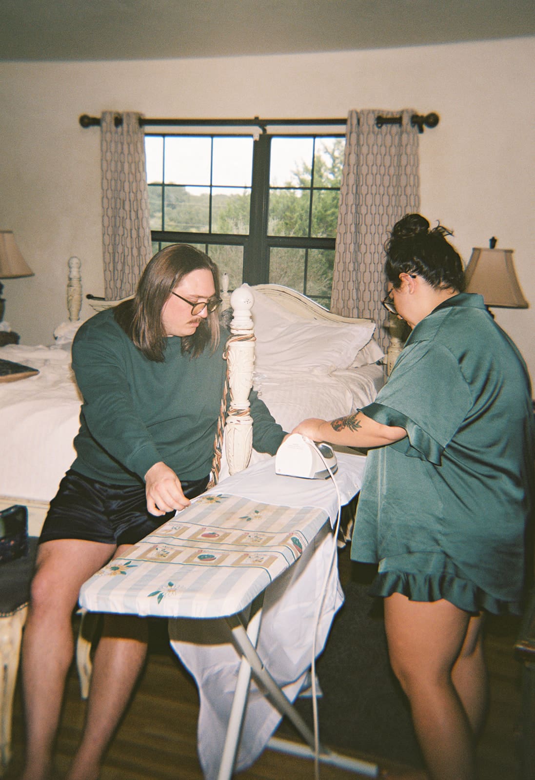 Two people stand at an ironing board in a bedroom; one is ironing fabric while the other arranges items on the board.